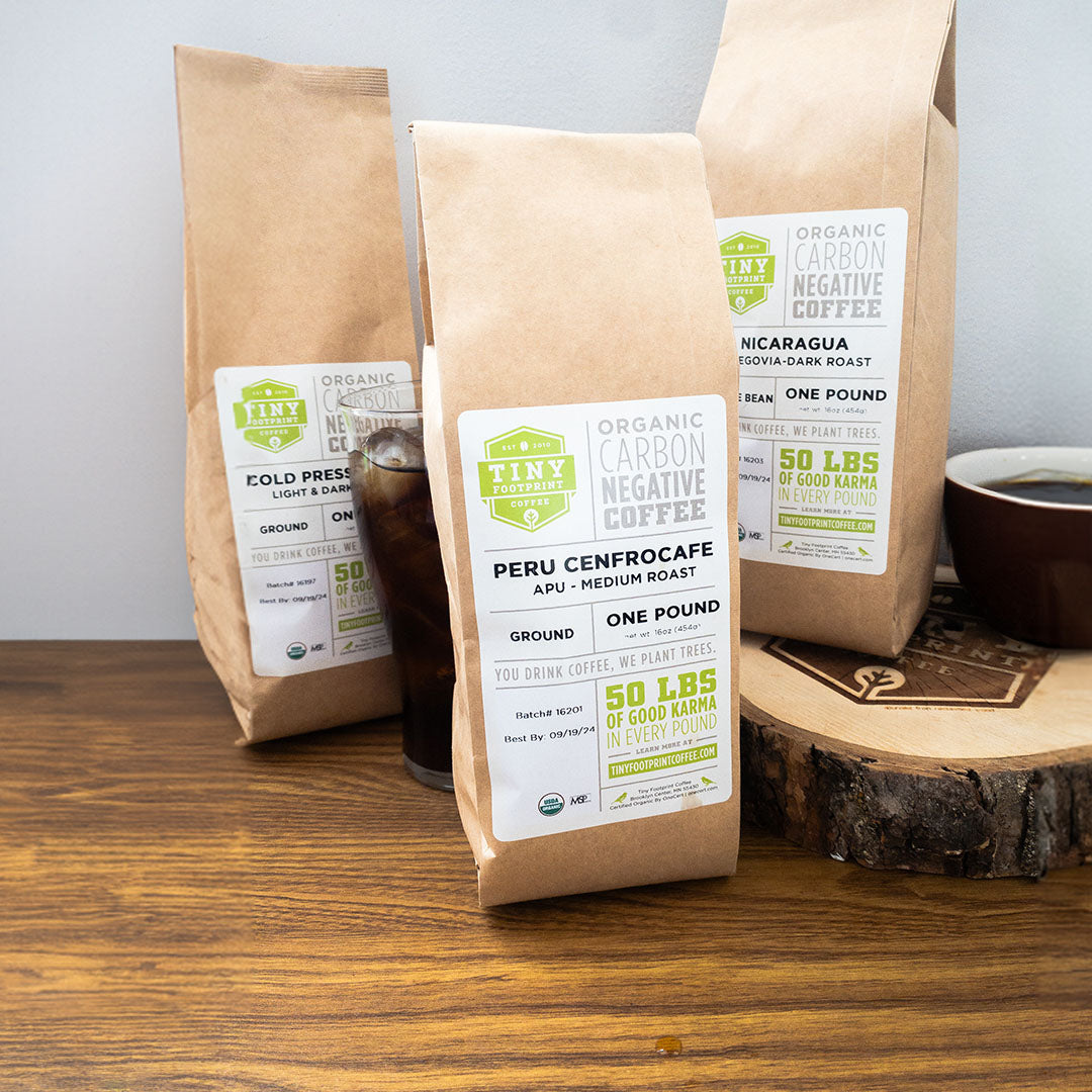 Three brown paper bags of Tiny Farm Coffee on a wooden surface with a cup of coffee.